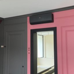 AIR Masters - Czarna klimatyzacja na różowej ścianie z dekoracyjnymi panelami w nowocześnie urządzonym wnętrzu z szarymi elementami i wzorzystą tapetą.