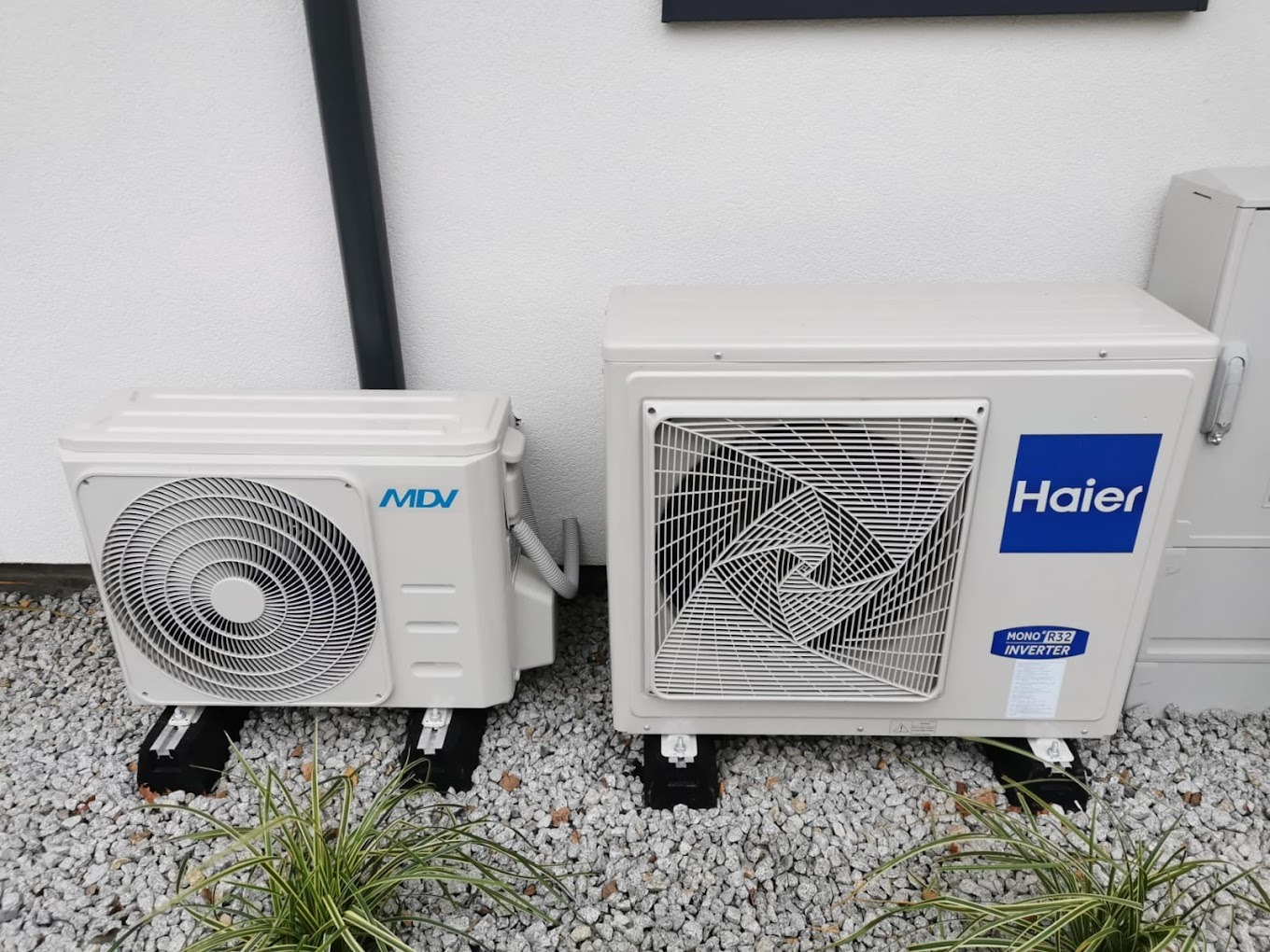 Dwa klimatyzatory: mniejszy MDV i większy Haier, ustawione na czarnych podstawkach na żwirowym podłożu, z zieloną rośliną w dolnej części kadru i białą ścianą w tle.