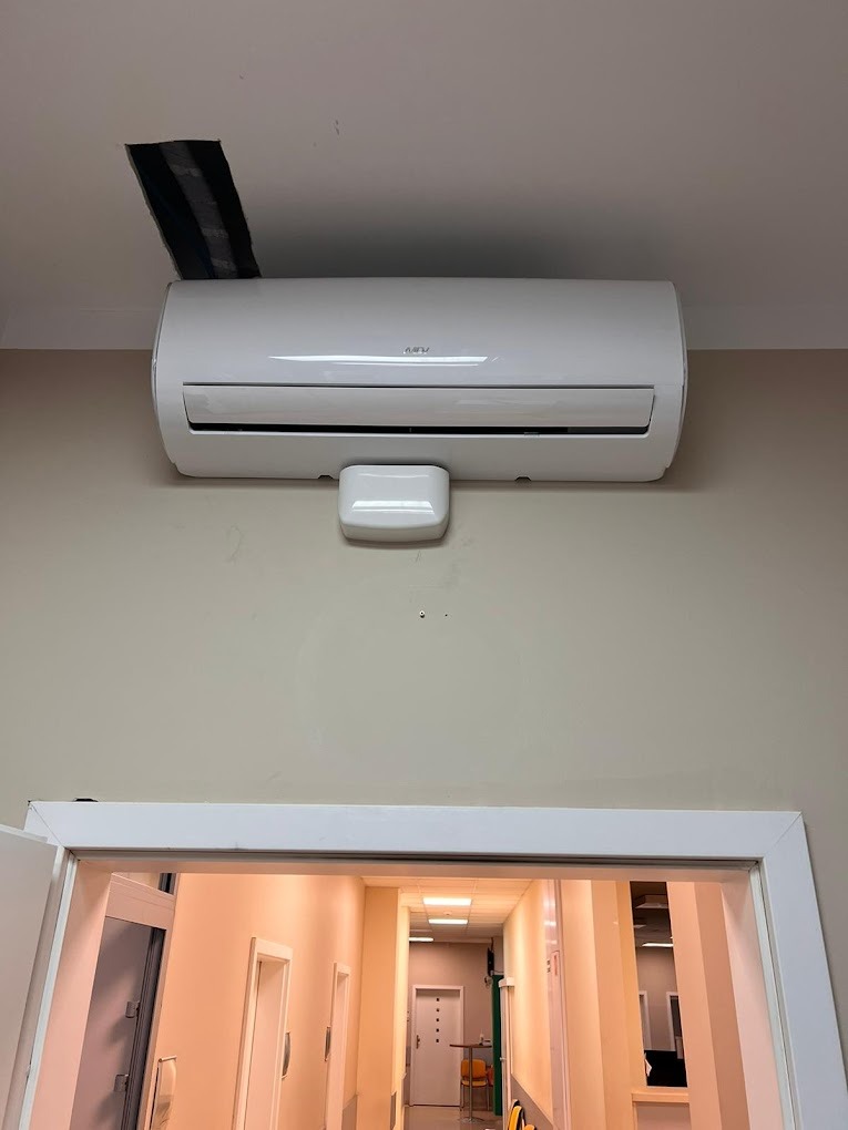 Biały klimatyzator ścienny w korytarzu, z widocznym fragmentem instalacji w suficie i perspektywą wnętrza przez otwarte drzwi.