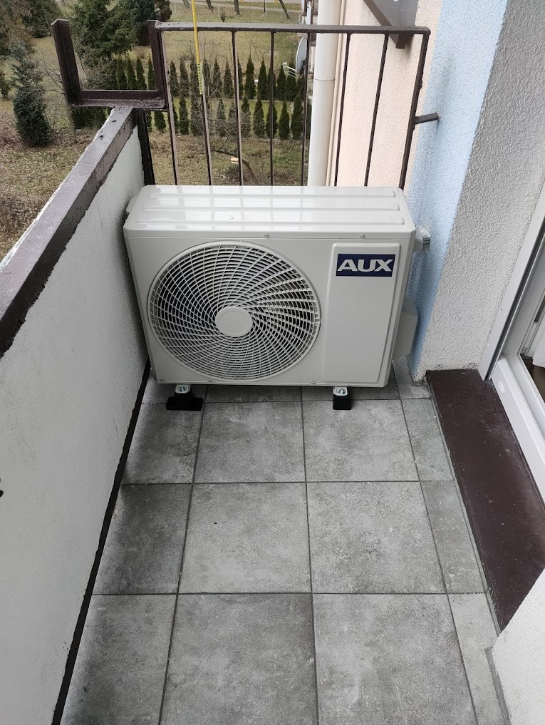 Biała jednostka zewnętrzna klimatyzacji AUX zamontowana na balkonie z szarymi płytkami. Widok z góry, balustrada i budynki w tle.