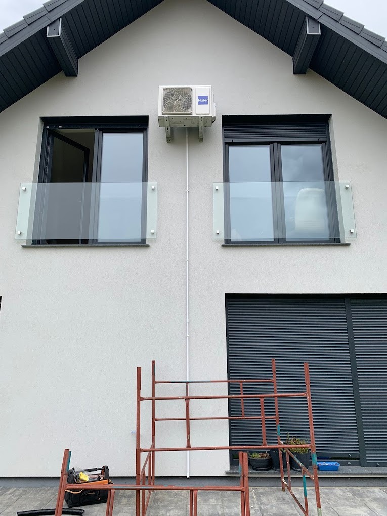 Biała jednostka klimatyzacji Haier zamontowana na elewacji domu, widoczne rury instalacyjne. Pod spodem rusztowanie i narzędzia.