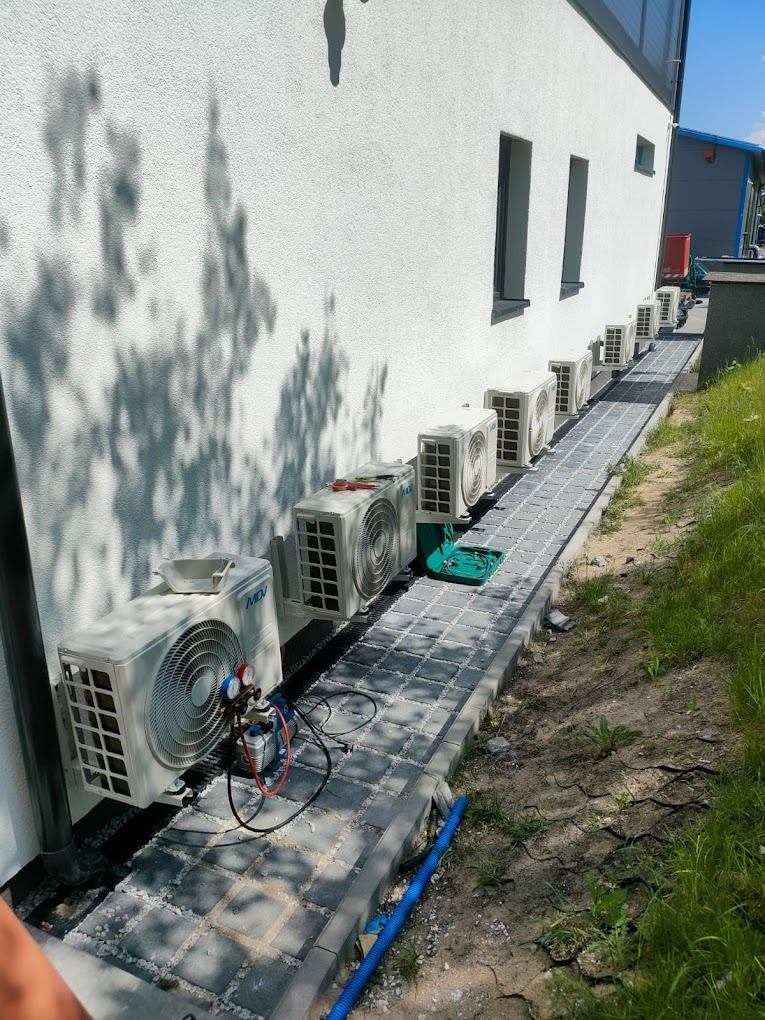 Rząd białych klimatyzatorów MDI zamontowanych na ścianie budynku, widoczne narzędzia serwisowe przy jednym z urządzeń, chodnik z kostki brukowej.