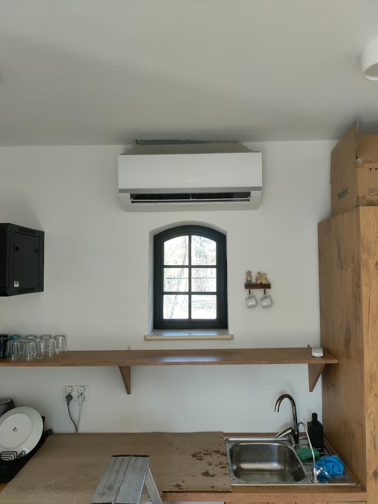 Biały klimatyzator ścienny nad oknem w kuchni z drewnianym blatem i zlewem. Widoczna drabina i naczynia. Minimalistyczny design, jasne wnętrze.