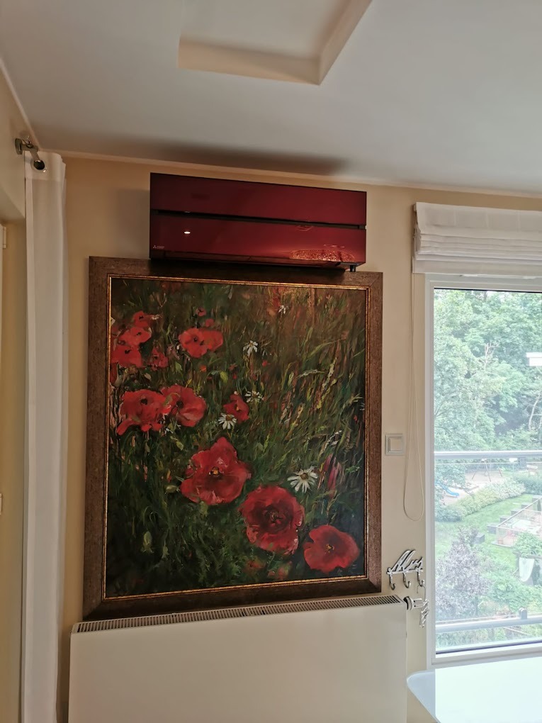 Czerwony klimatyzator ścienny nad obrazem z motywem maków w ozdobnej ramie, w jasnym wnętrzu z oknem i grzejnikiem.