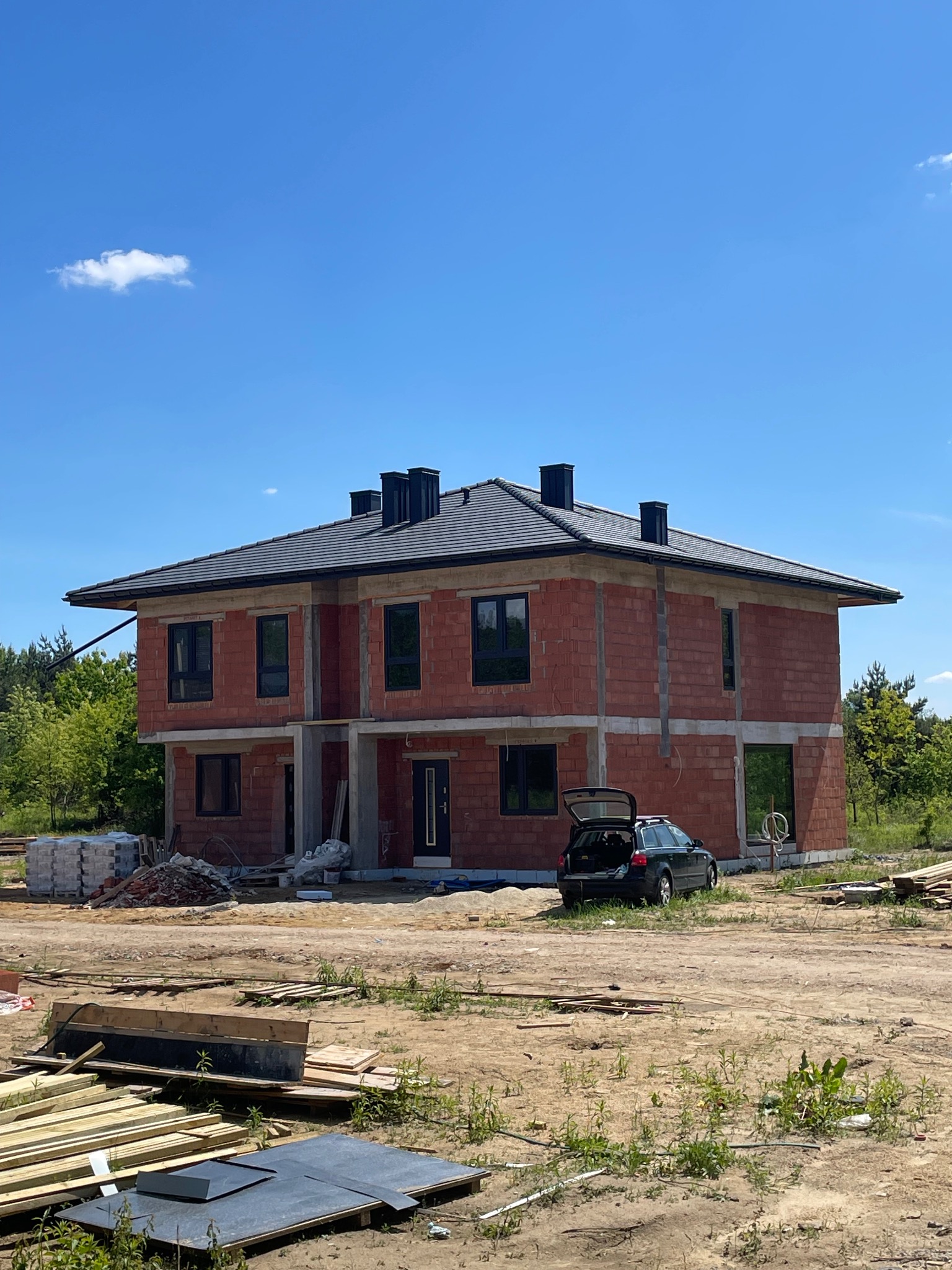 Nowoczesny dom w budowie z ciemną dachówką w Wieliszewie, widok z zewnątrz. Dach pokryty, kominy zamontowane, okna wstawione.