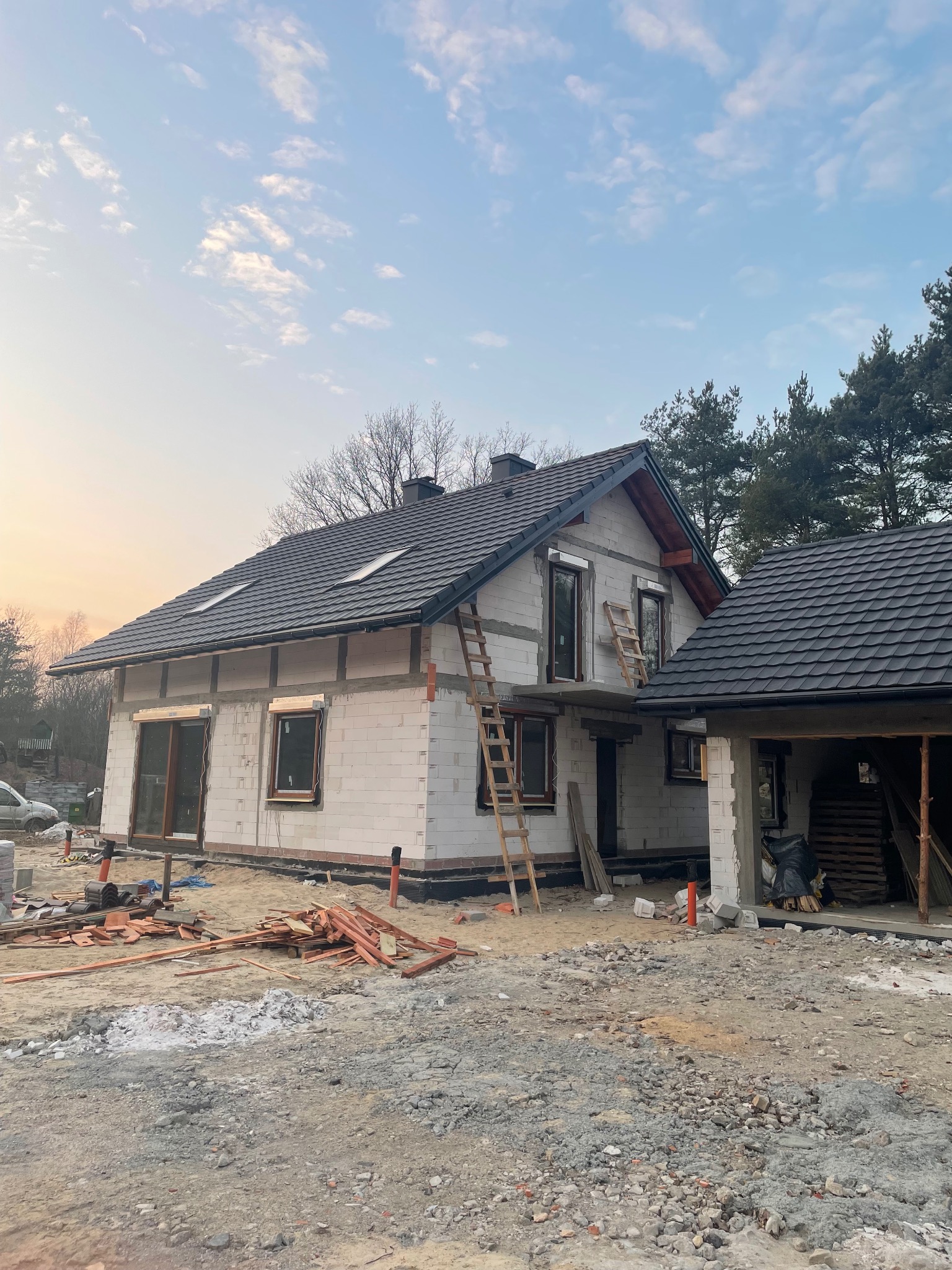 Dom jednorodzinny w trakcie budowy, z ciemną dachówką i oknami. Widoczne rusztowania i materiały budowlane. Ujęcie z poziomu gruntu, w tle drzewa i błękitne niebo.