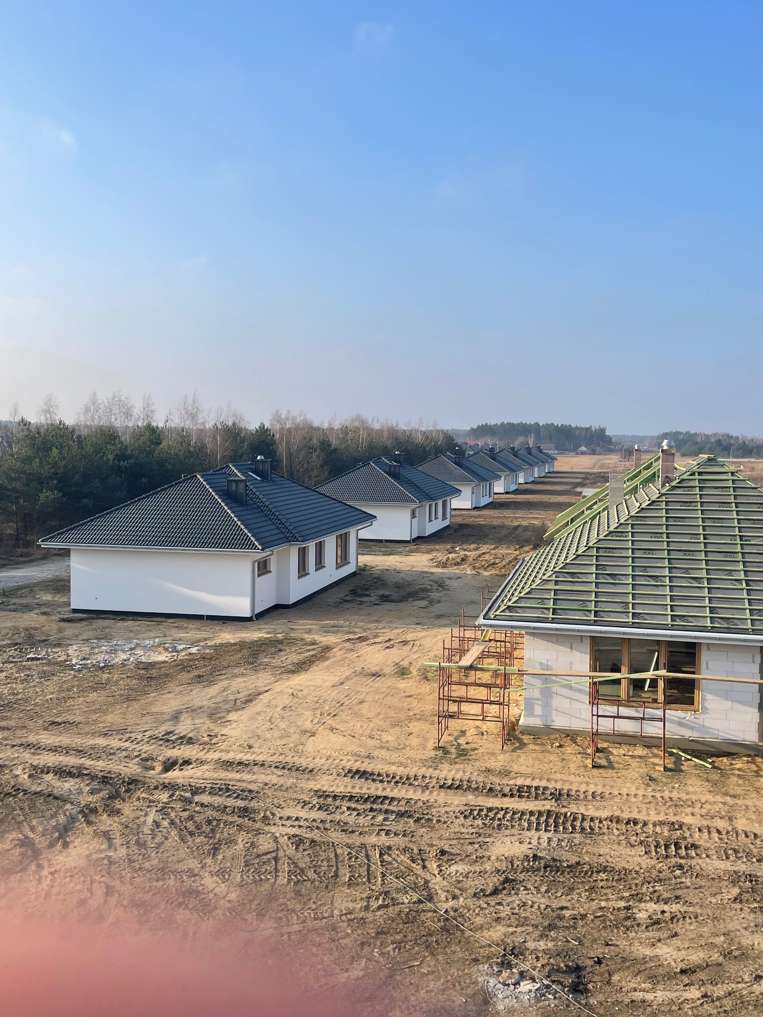 Szereg identycznych domów jednorodzinnych w trakcie budowy, z dachem w różnym stadium zaawansowania, od konstrukcji po gotowe pokrycie z czarnej dachówki.