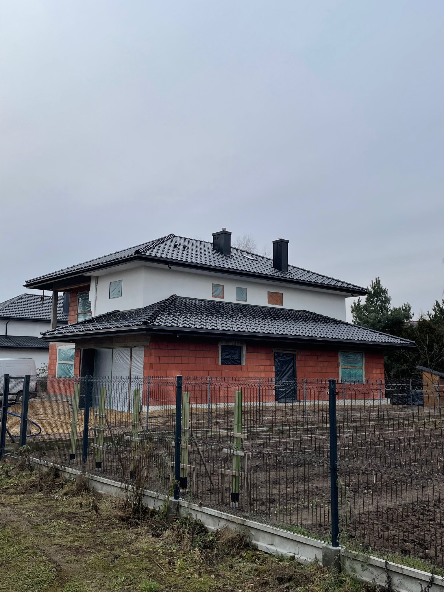 Nowoczesny dom w trakcie budowy z czarnym dachem z dachówki ceramicznej. Widoczne kominy i ogrodzenie panelowe. Budynek dwukondygnacyjny z elementami w stanie surowym.