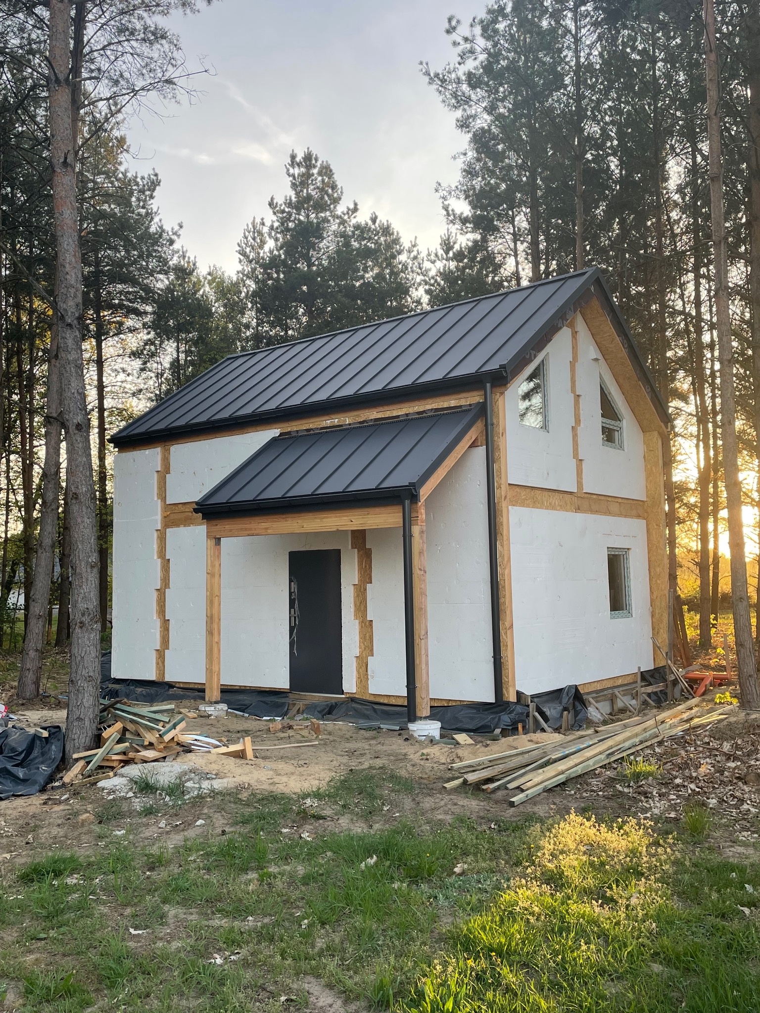 Dom w trakcie budowy z czarnym dachem i białymi ścianami, otoczony drzewami. Widoczne elementy konstrukcyjne i materiały budowlane. Częściowo ocieplony budynek.