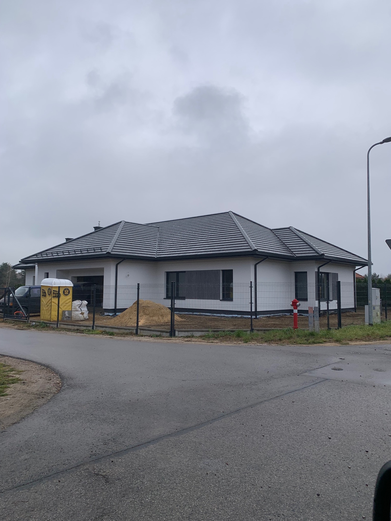 Nowoczesny dom jednorodzinny z ciemną dachówką karpiówką i białymi ścianami, ogrodzony siatką. Widoczne materiały budowlane i hydrant. Pochmurne niebo w tle.