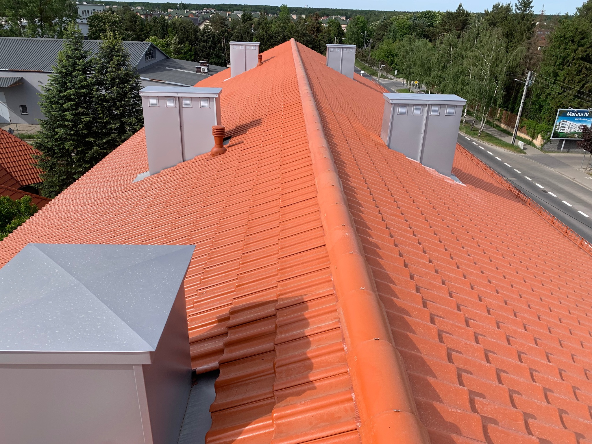 Czerwony dach z dachówki ceramicznej, widok z góry, z czterema kominami z szarej blachy. W tle drzewa i budynki.