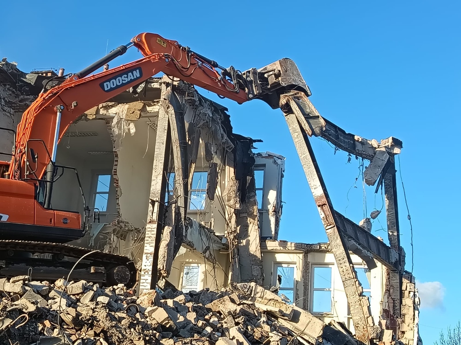 Pomarańczowa koparka Doosan z hydraulicznymi nożycami rozbiera budynek z białej cegły, z widocznymi oknami, na tle błękitnego nieba. Wokół gruz.