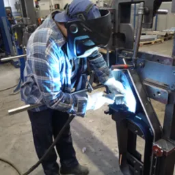 Spawacz w masce, w niebieskiej kraciastej koszuli, łączy metalowe elementy. Iskry i jasne światło spawania. Tło to hala przemysłowa z maszynami.