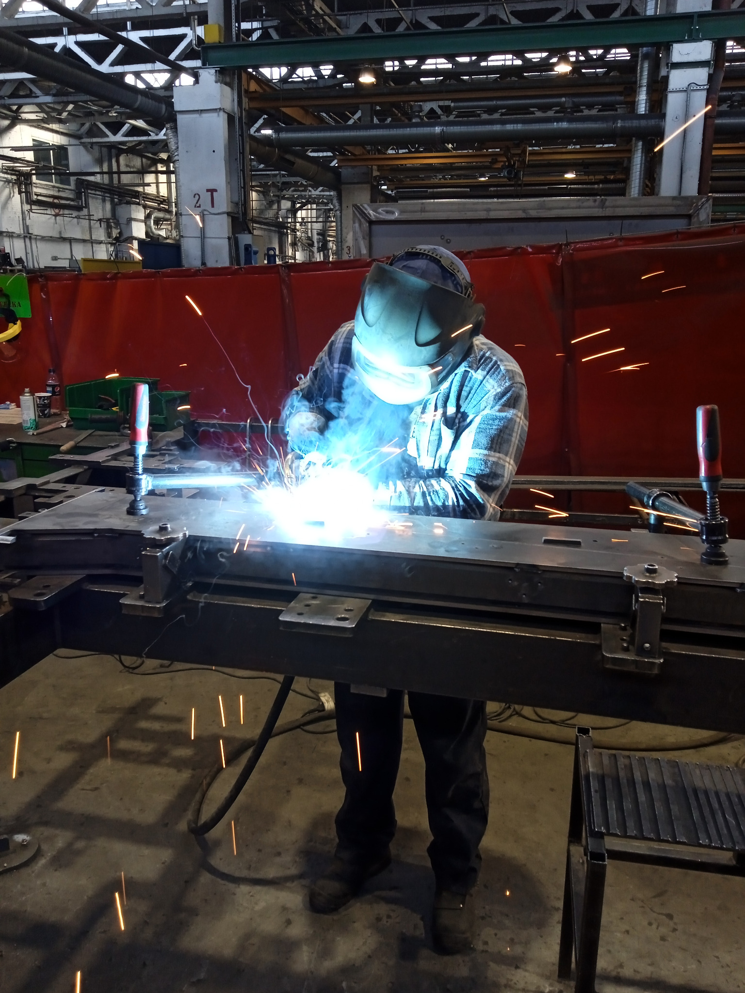 Pracownik w masce spawalniczej łączy metalowe elementy w hali produkcyjnej. Widoczne iskry i intensywne światło procesu spawania. W tle konstrukcja hali.