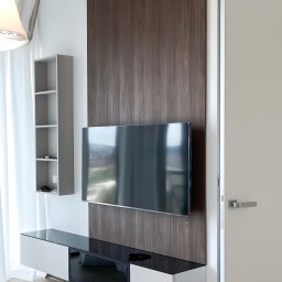 ANNA BUCZNY PIĘKNE WNĘTRZA - Nowoczesny salon z telewizorem na tle drewnianej ściany, szafka RTV z czarnym blatem, półka wisząca. Minimalistyczny design, jasne wnętrze, ukryte drzwi.