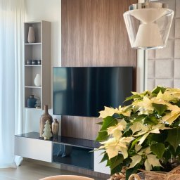 ANNA BUCZNY PIĘKNE WNĘTRZA - Nowoczesny salon z telewizorem na drewnianej ścianie, białą szafką RTV, półką z dekoracjami i lampą wiszącą. Wnętrze doświetlone naturalnym światłem.