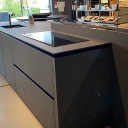 ANNA BUCZNY PIĘKNE WNĘTRZA - Nowoczesna wyspa kuchenna z szarym blatem i minimalistycznym zlewem oraz płytą indukcyjną. W tle regał z książkami i dekoracjami. Stonowana kolorystyka, elegancki design.