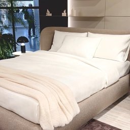 ANNA BUCZNY PIĘKNE WNĘTRZA - Beżowe łóżko z białymi poduszkami i narzutą w minimalistycznej sypialni z półkami dekoracyjnymi i widokiem na zieleń za oknem.