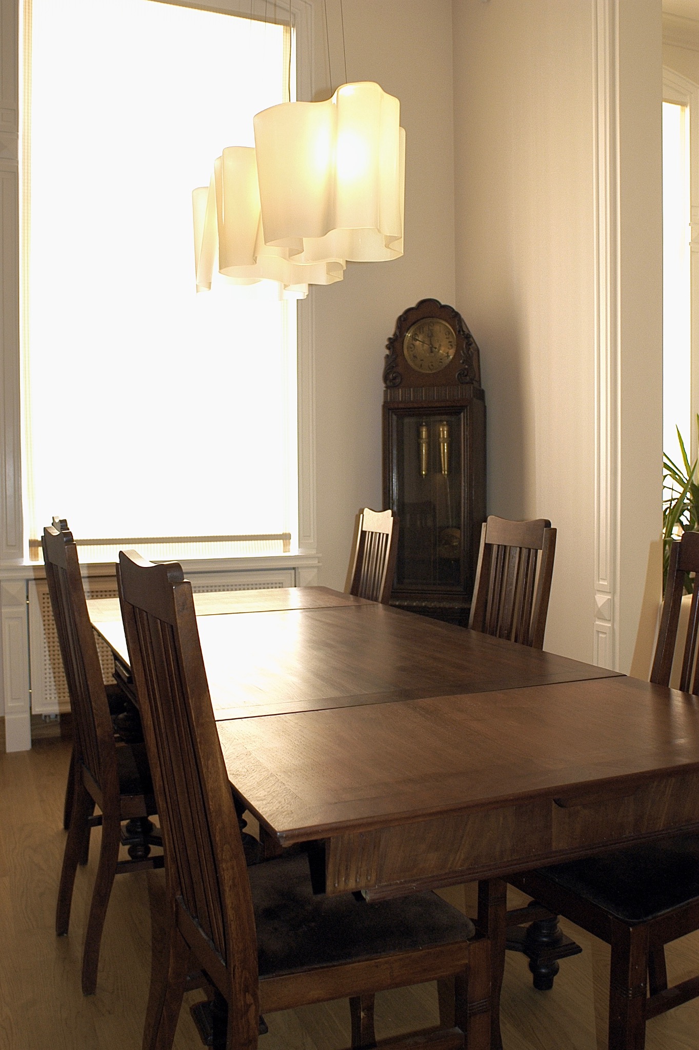 Jadalnia w stylu vintage: drewniany stół z wysuwaną szufladą, krzesła z wysokimi oparciami, zegar stojący i designerska lampa wisząca.