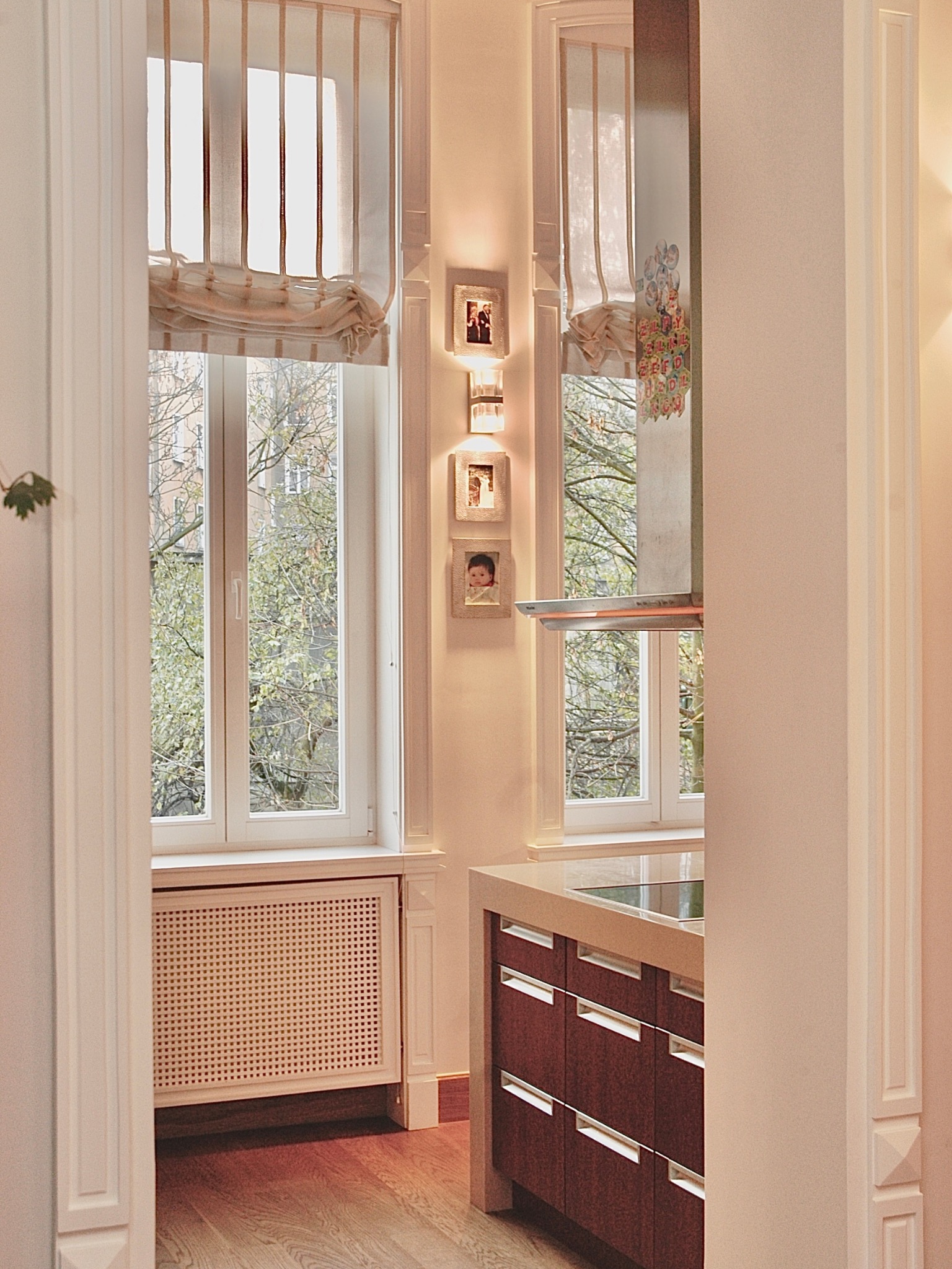 Eklektyczna kuchnia z półwyspem: drewniane szafki, jasny blat, okna z roletami rzymskimi i galeria zdjęć na ścianie w kamienicy. Widok z perspektywy wejścia.