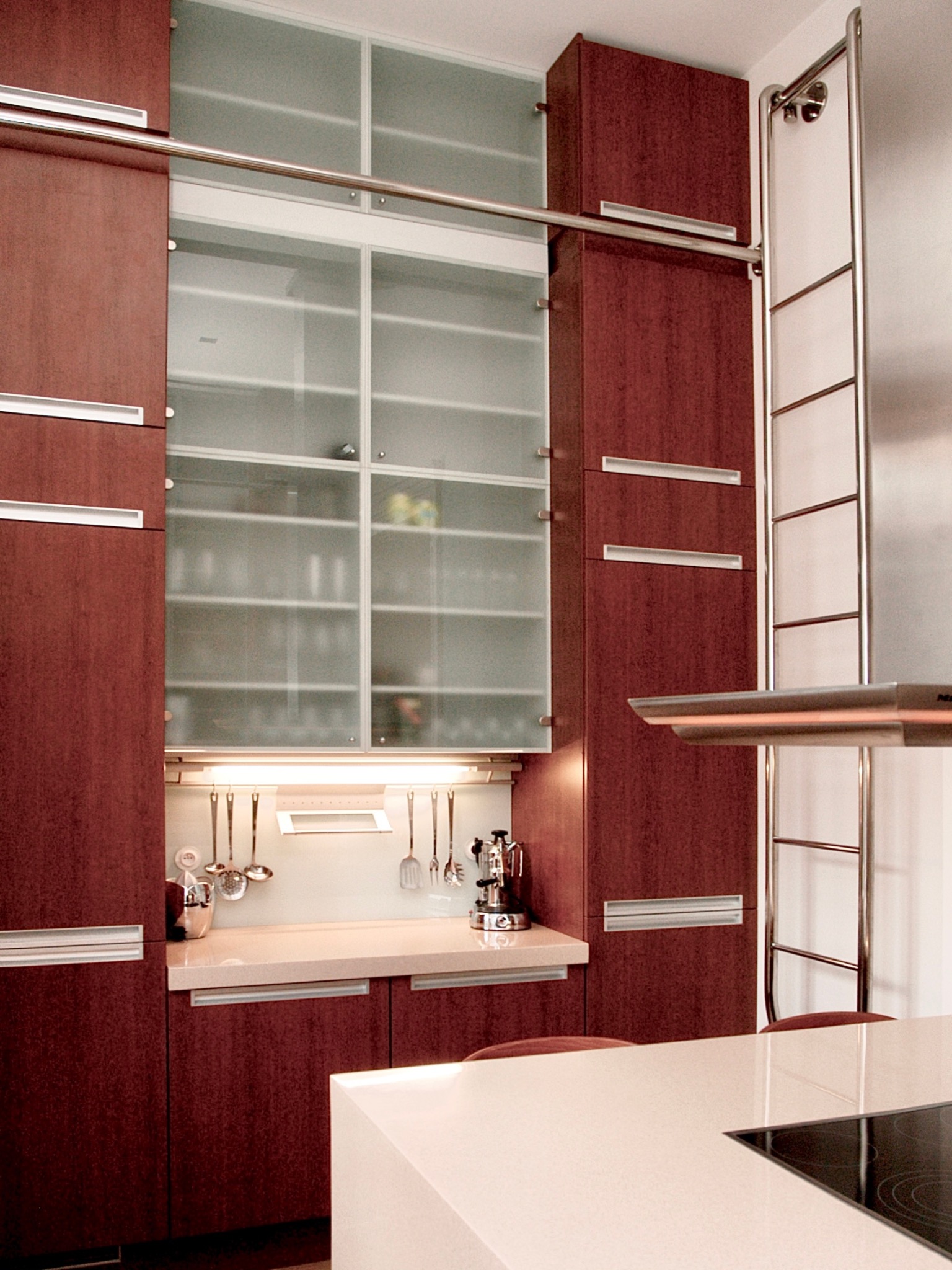 Ciemna, nowoczesna kuchnia z zabudową do sufitu. Szklane witryny, metalowe akcenty i oświetlenie podszafkowe. Widoczny fragment wyspy kuchennej z płytą.