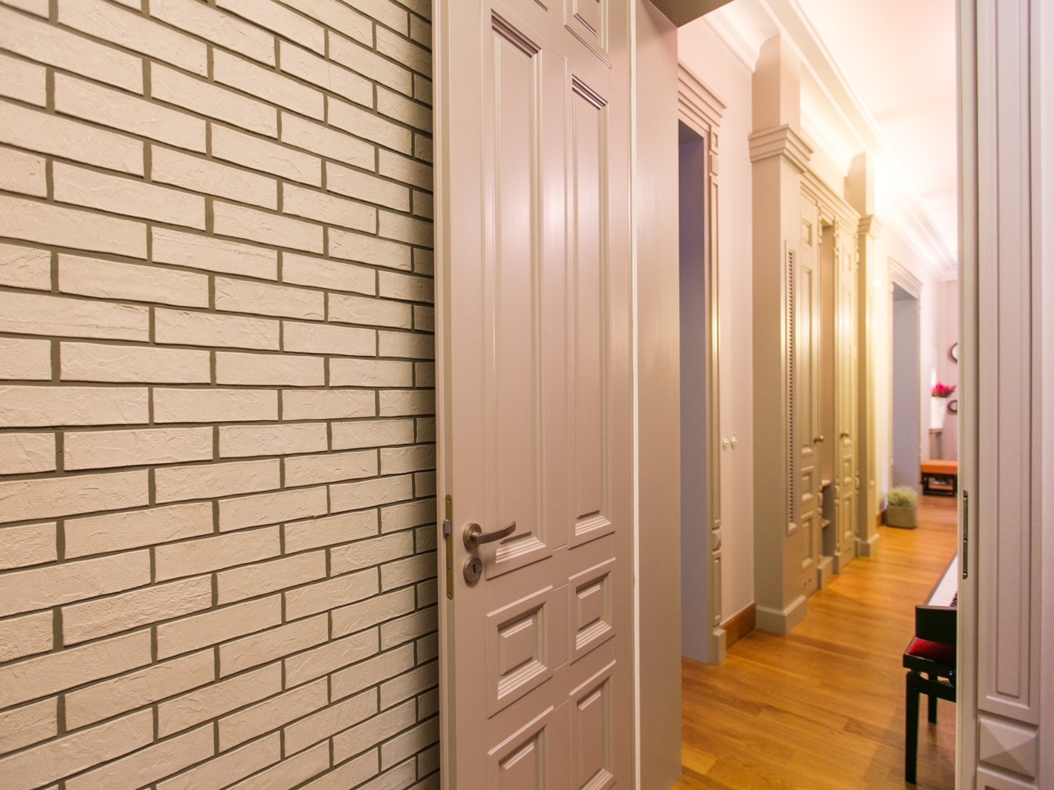 Korytarz w stylu vintage: ceglana ściana, otwarta, kremowa  drewniana  rama drzwiowa z ozdobnymi panelami i widok na wnętrze z zabudową i drewnianą podłogą.