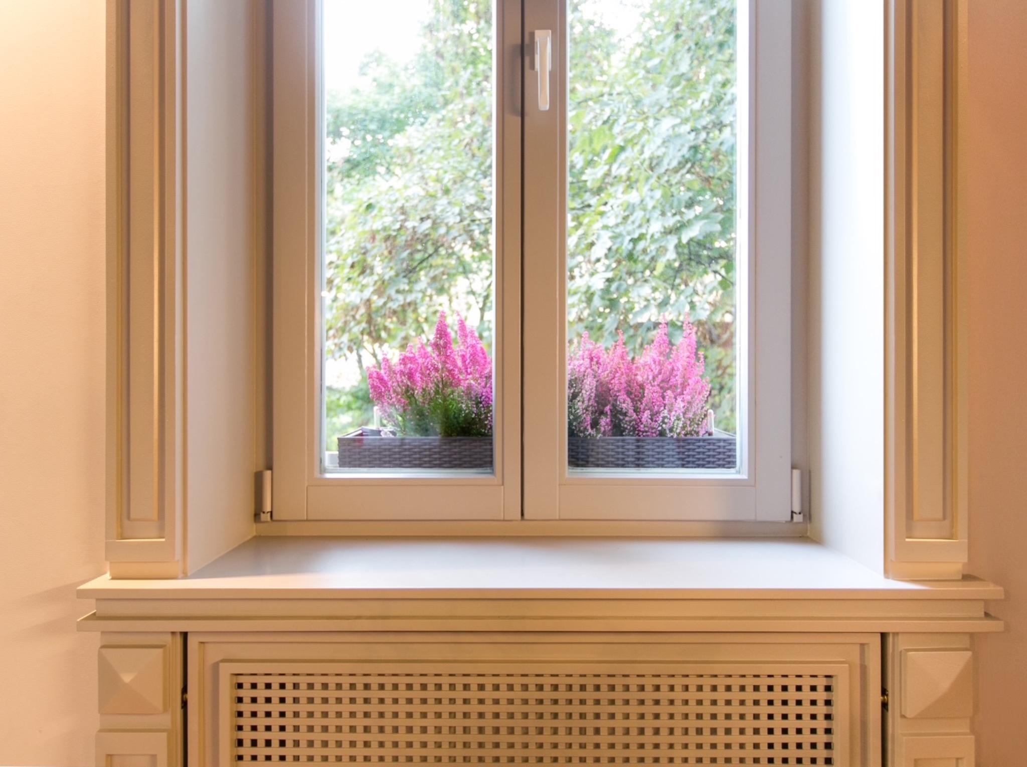 Biała zabudowa okna z grzejnikiem, dekoracyjne panele, różowe wrzosy w doniczkach na parapecie, widok na zieleń za oknem. Klasyczny styl, jasne wnętrze.