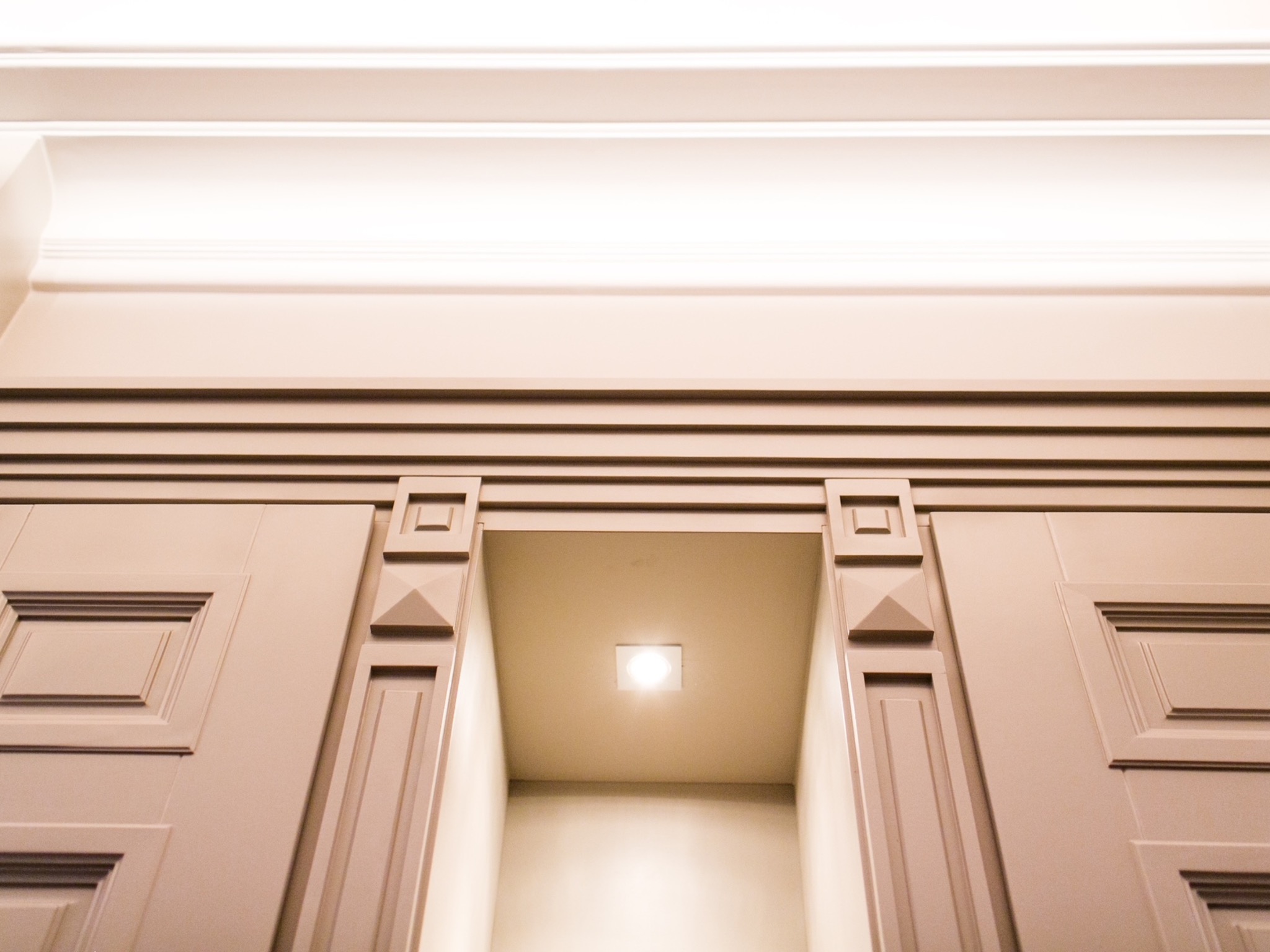 Detal zabudowy meblowej w odcieniach beżu z oświetleniem punktowym. Klasyczna stolarka z dekoracyjnymi frezami i geometrycznymi zdobieniami. Ujęcie z dołu.