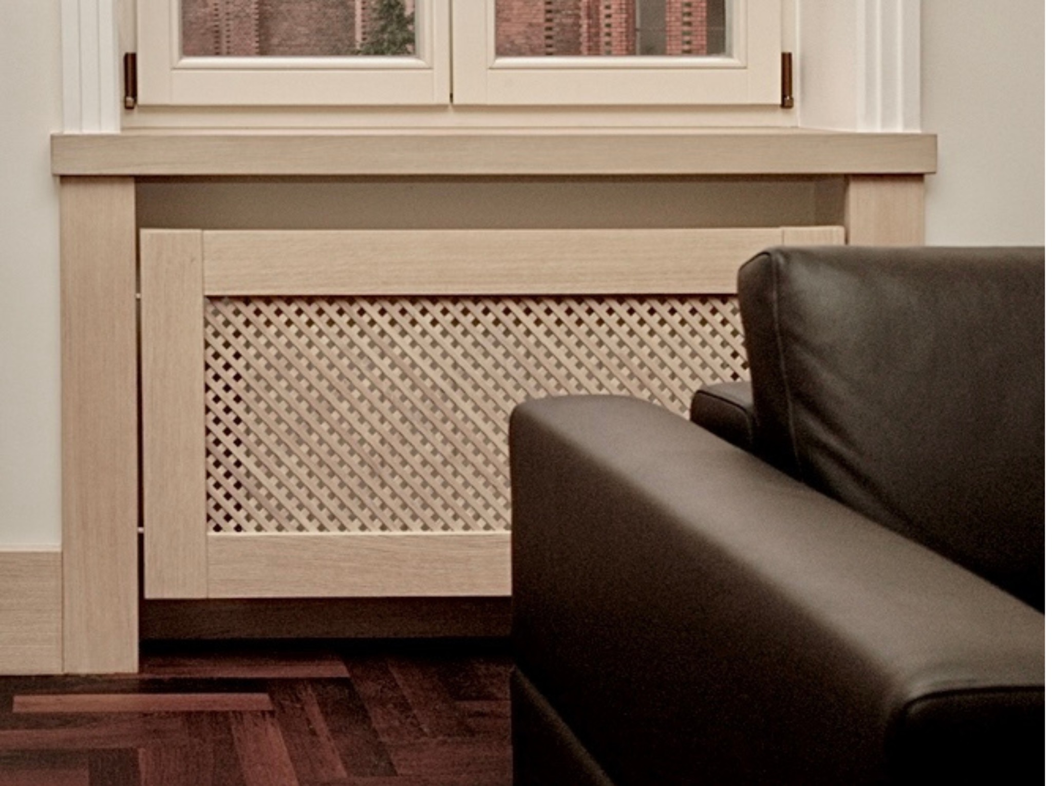 Zabudowa grzejnika z drewnianą kratką w salonie, dopasowana do ramy okiennej. Widoczny fragment skórzanego fotela i parkiet. Styl vintage, eklektyczna kamienica.