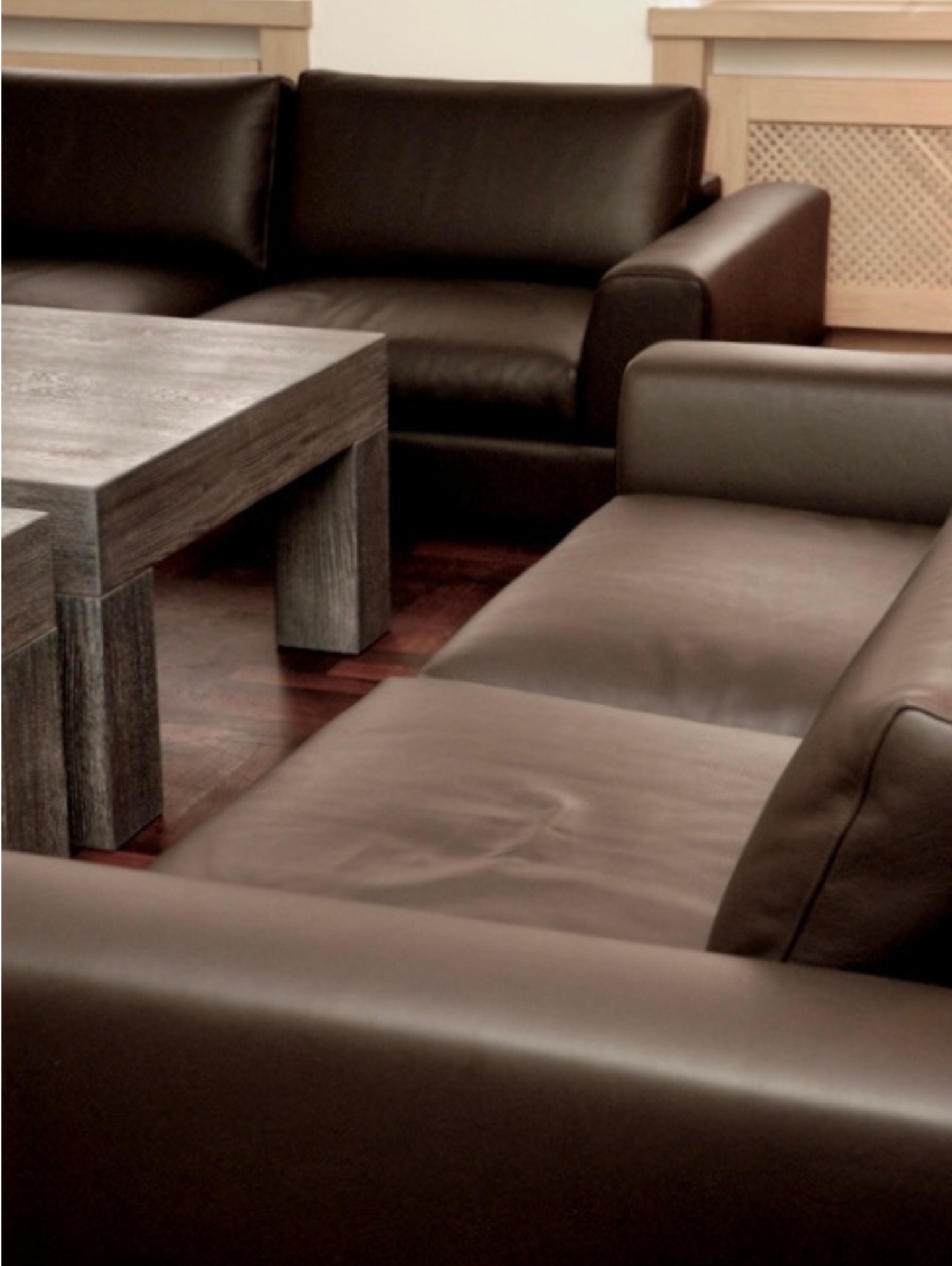 Ciemnobrązowy, skórzany narożnik z drewnianym stolikiem kawowym na parkiecie. W tle zabudowa grzejnika. Minimalistyczny salon w neutralnej tonacji.