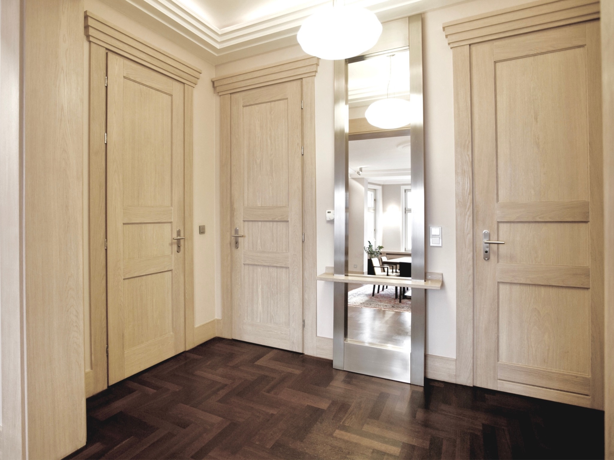 Hol z parkietem w jodełkę, trzy drewniane drzwi z dekoracyjnymi listwami, lustro w metalowej ramie odbijające wnętrze salonu z lampą i stołem.