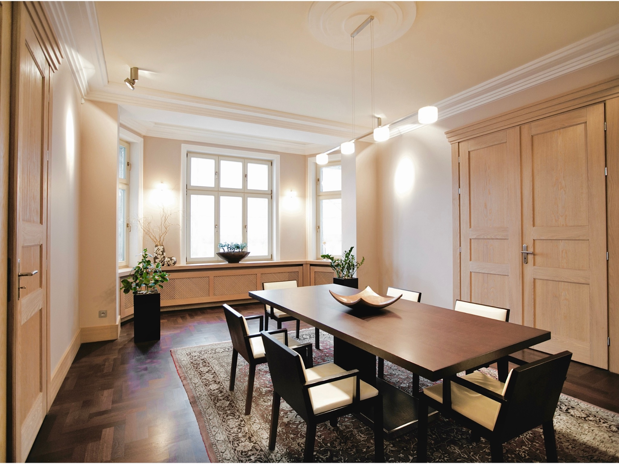 Salon w kamienicy: ciemna podłoga z drewna, jasne ściany, jesionowa stolarka. Stół z krzesłami na dywanie, oświetlenie sufitowe, rośliny w donicach.