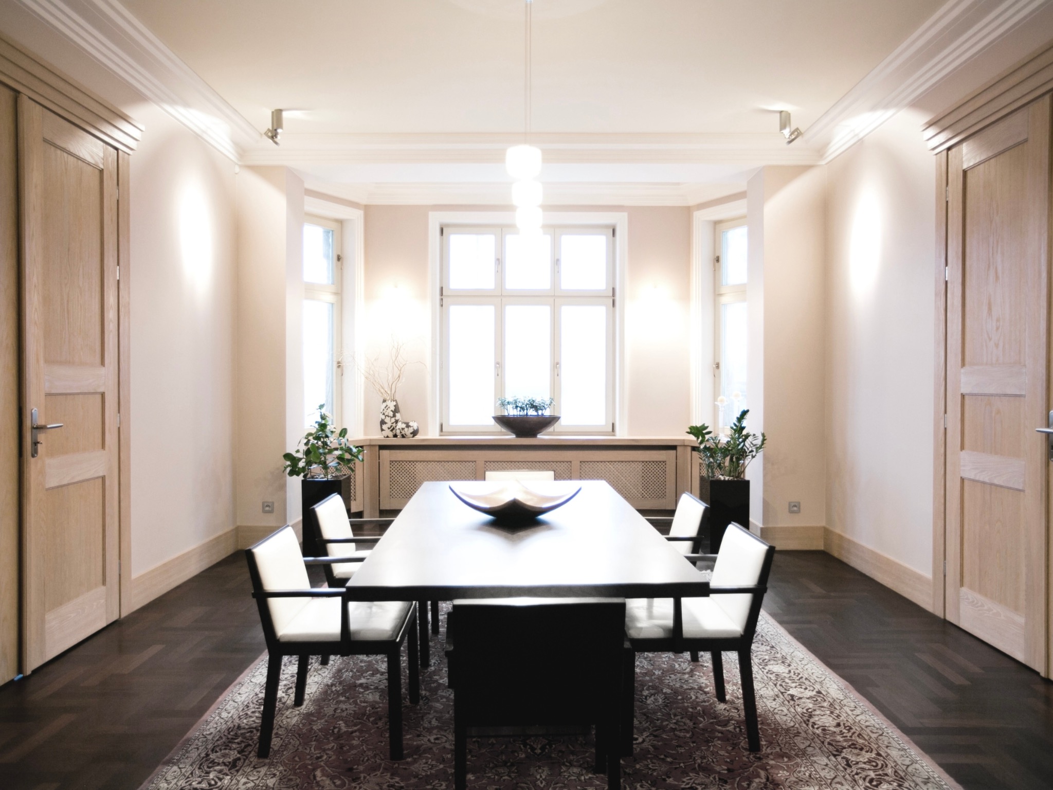 Elegancka jadalnia z ciemnym stołem i jasnymi krzesłami, na dywanie w orientalny wzór. Jasne ściany, drewniane drzwi i okno z roślinami dodają wnętrzu klasy.