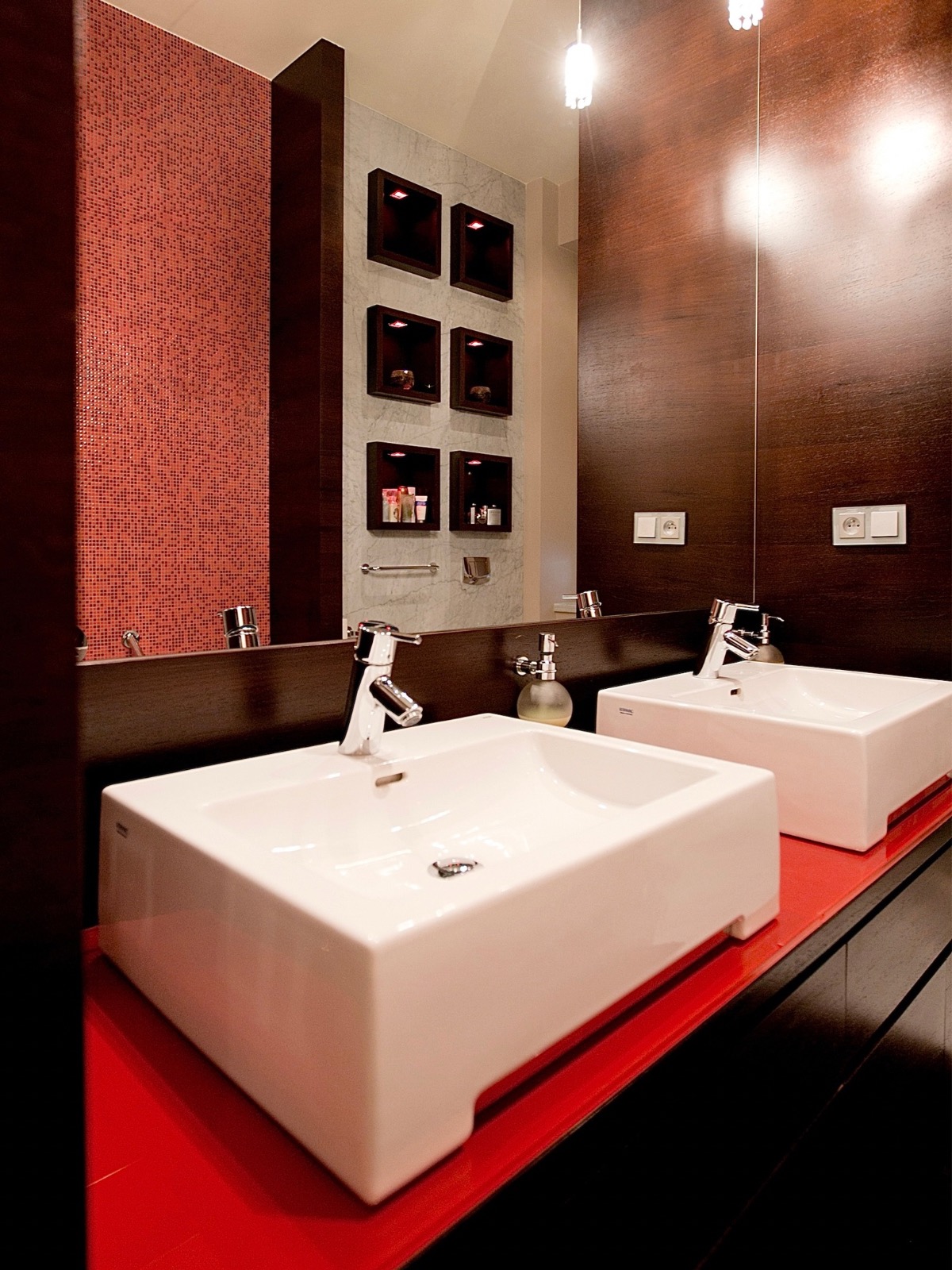 Nowoczesna łazienka z dwoma białymi umywalkami na czerwonym blacie, drewnianą ścianą i mozaiką. W tle półki z oświetleniem i marmurowa ściana.