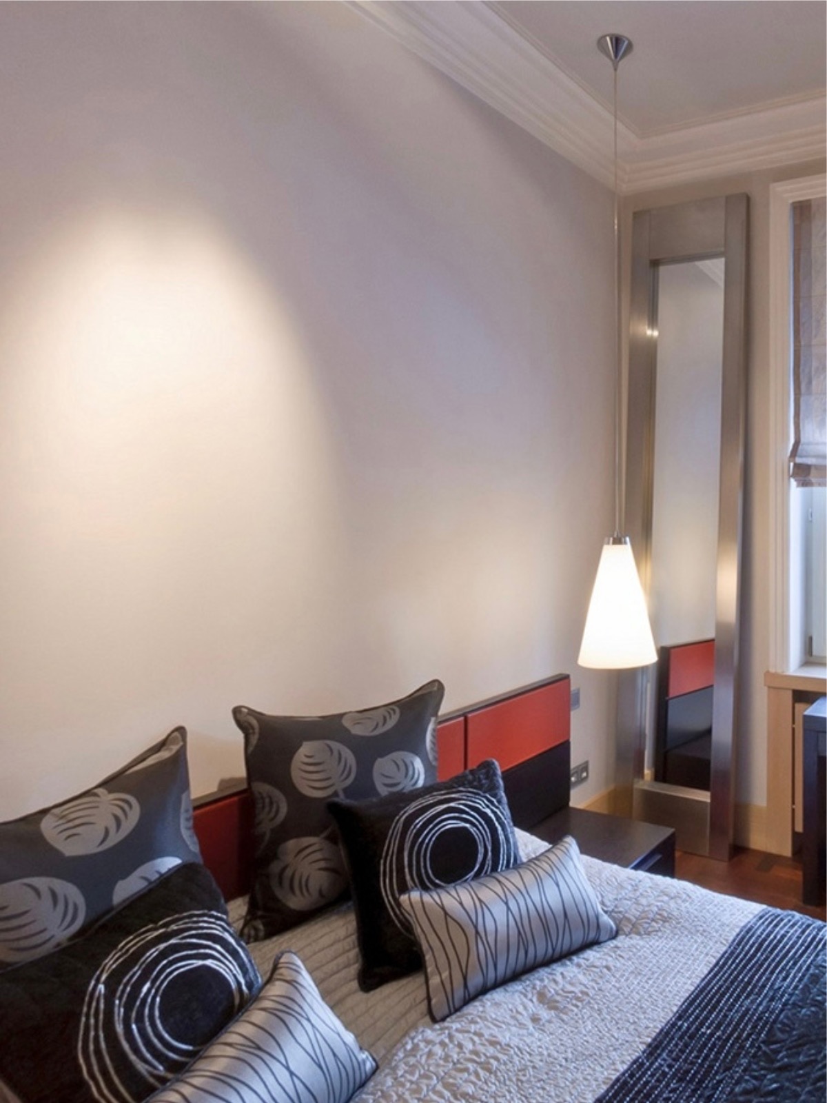 Nowoczesna sypialnia z łóżkiem, dekoracyjnymi poduszkami i wysokim lustrem w metalowej ramie. Nad łóżkiem wisi lampa z białym kloszem. Stonowana kolorystyka.