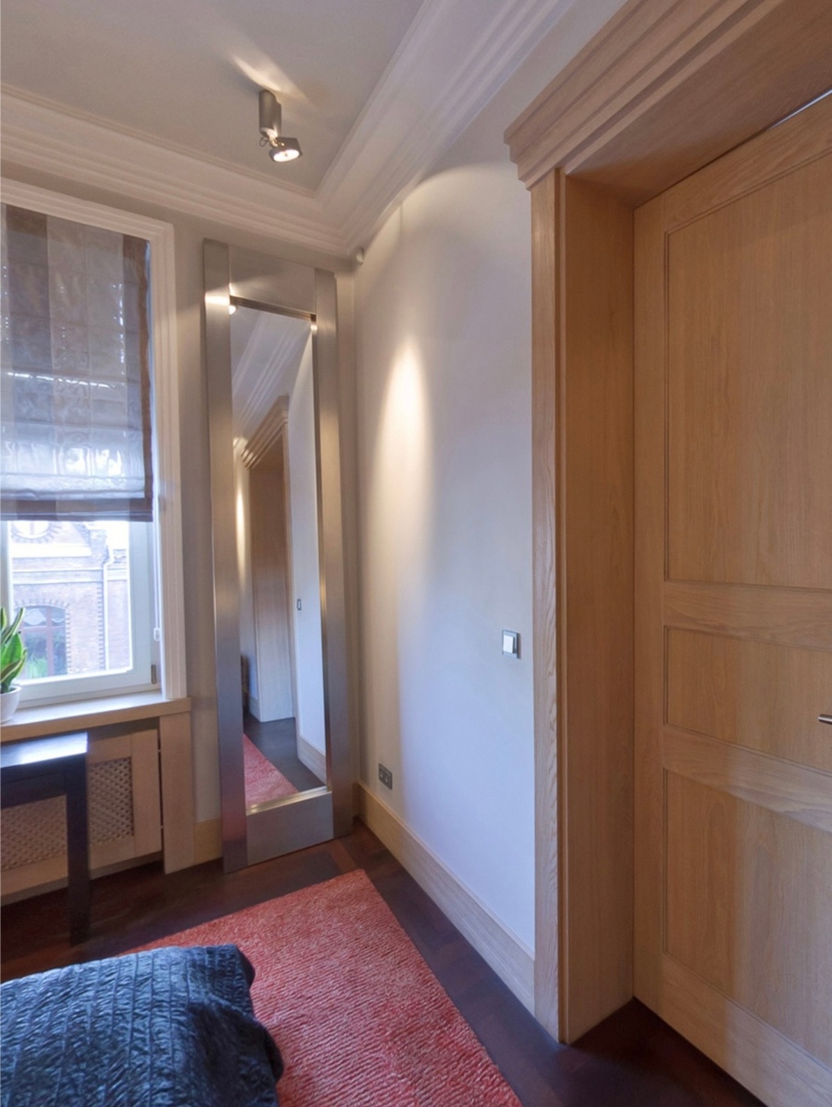 Nowoczesne wnętrze z lustrem w metalowej ramie, drewnianą stolarką drzwiową i oknem z żaluzjami. Widoczny fragment dywanu i łóżka.