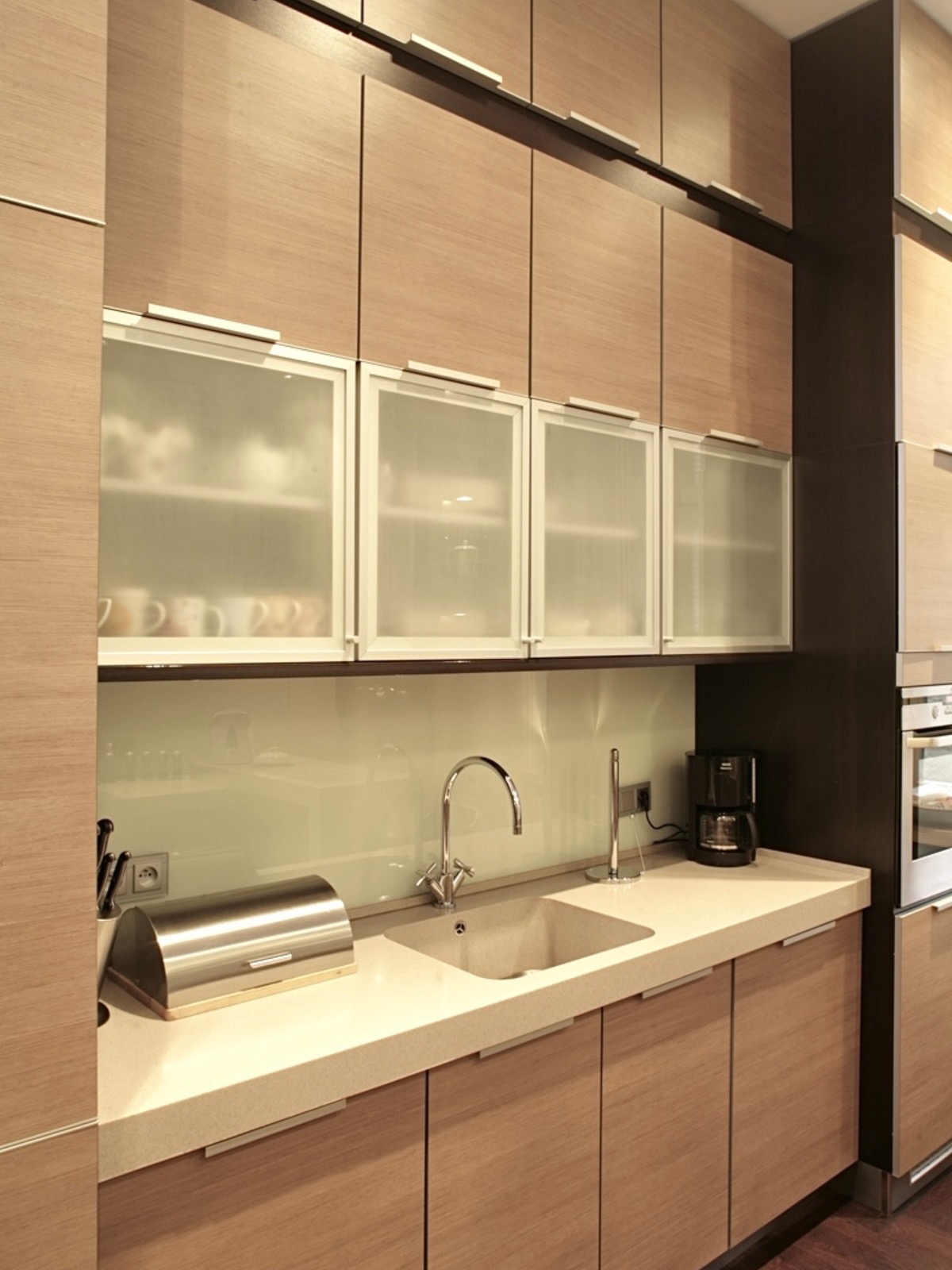 Jasna kuchnia z drewnianymi szafkami, przeszklonymi górnymi szafkami i beżowym blatem. Na blacie widoczny zlew, bateria, chlebak i ekspres do kawy.