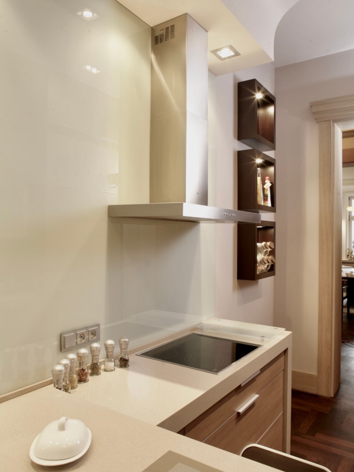 Nowoczesna kuchnia z płytą ceramiczną i pochłaniaczem. Na blacie przyprawy w młynkach. W tle otwarte półki z oświetleniem i widok na salon.