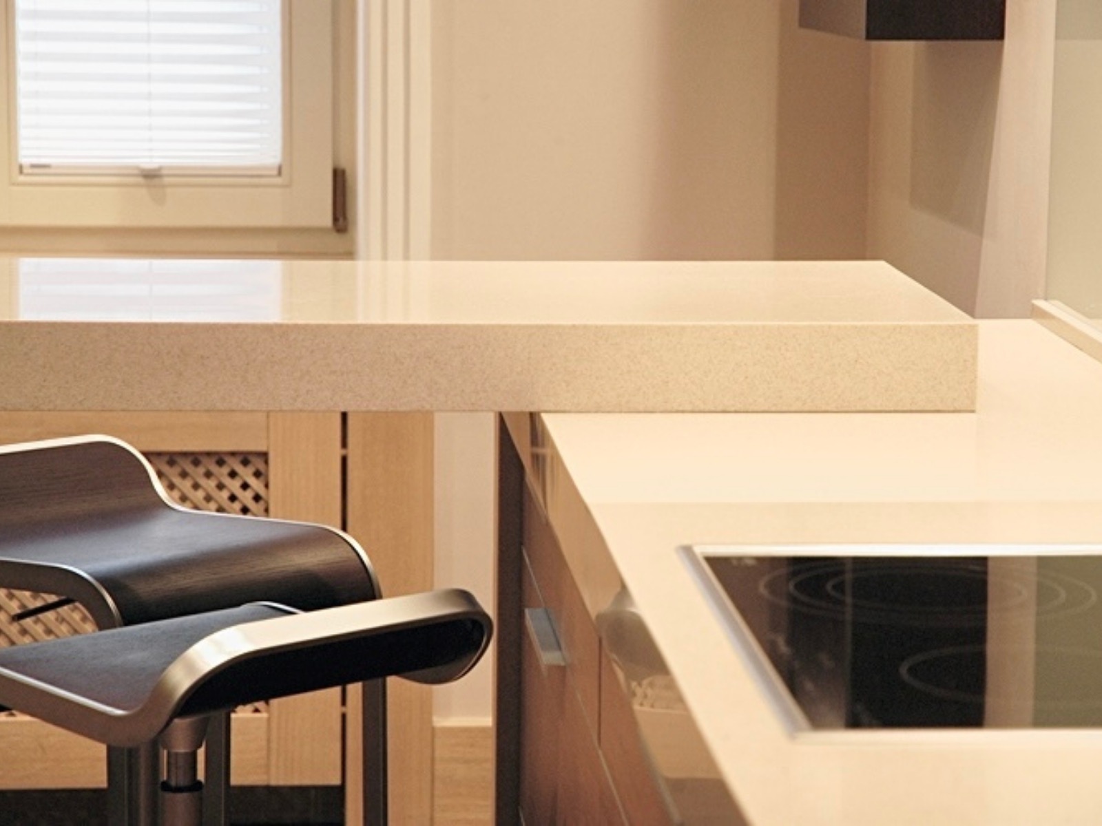 Beżowa kuchnia z blatem z konglomeratu i hokerami. Minimalistyczny design z płytą indukcyjną i oknem w tle. Ujęcie z niskiego kąta podkreśla detale.