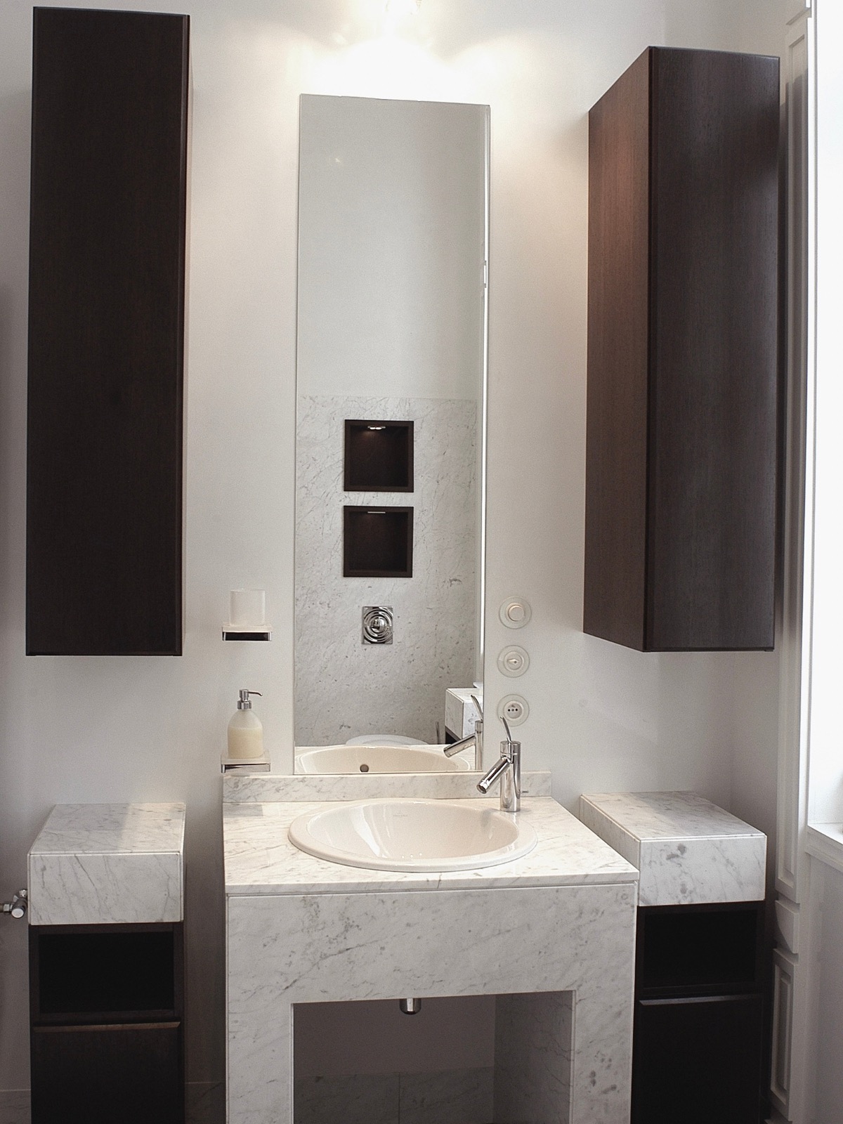 Nowoczesna łazienka z marmurową umywalką i ciemnymi, wiszącymi szafkami. Minimalistyczny design z lustrem i subtelnym oświetleniem punktowym.