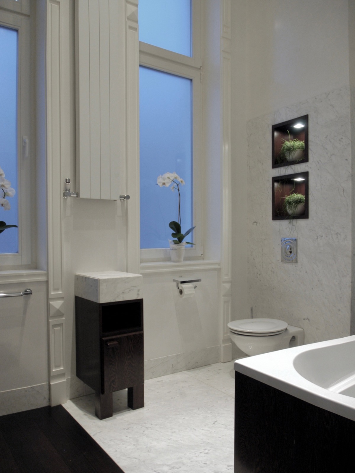 Jasna łazienka w stylu vintage z marmurową ścianą, toaletą, szafką z umywalką i roślinami w podświetlanych niszach. W oknach widoczne kwiaty.