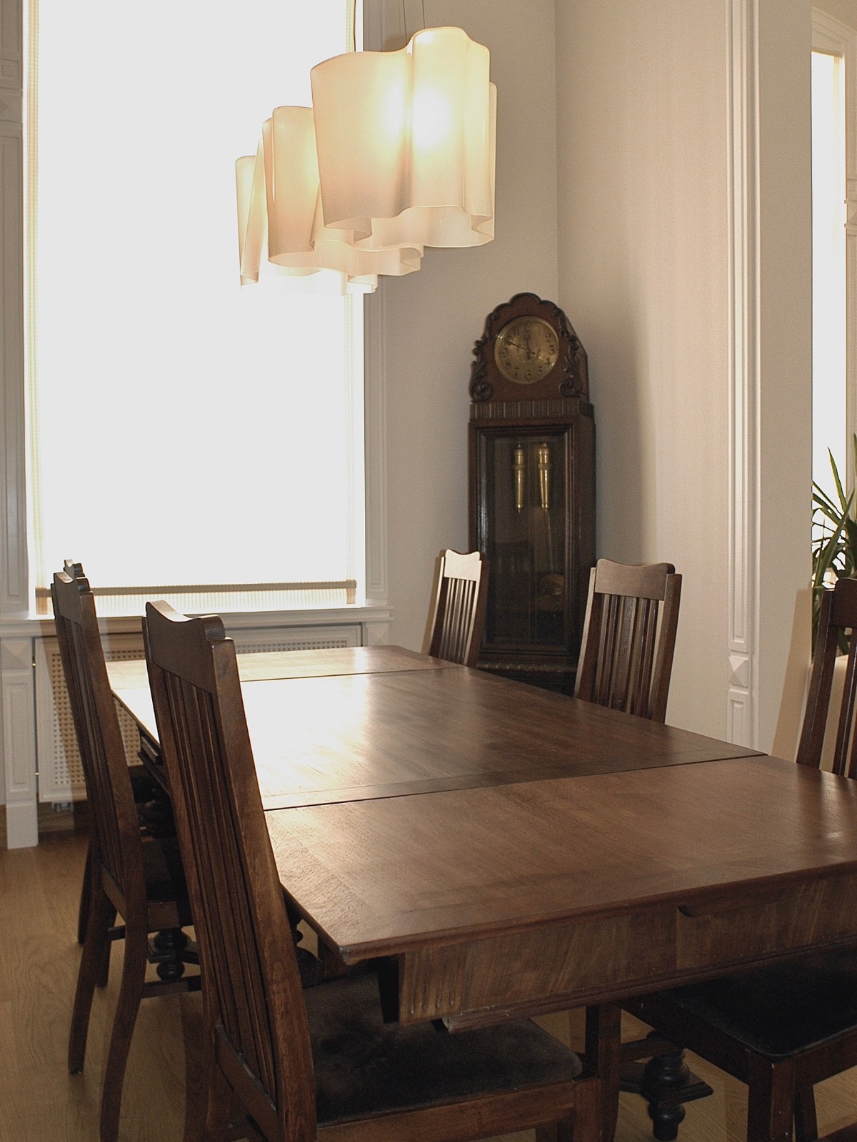 Jadalnia w kamienicy: drewniany stół z krzesłami, zegar stojący i lampa wisząca. Kontrast bieli ścian z ciemnym wyposażeniem. Ujęcie z perspektywy.