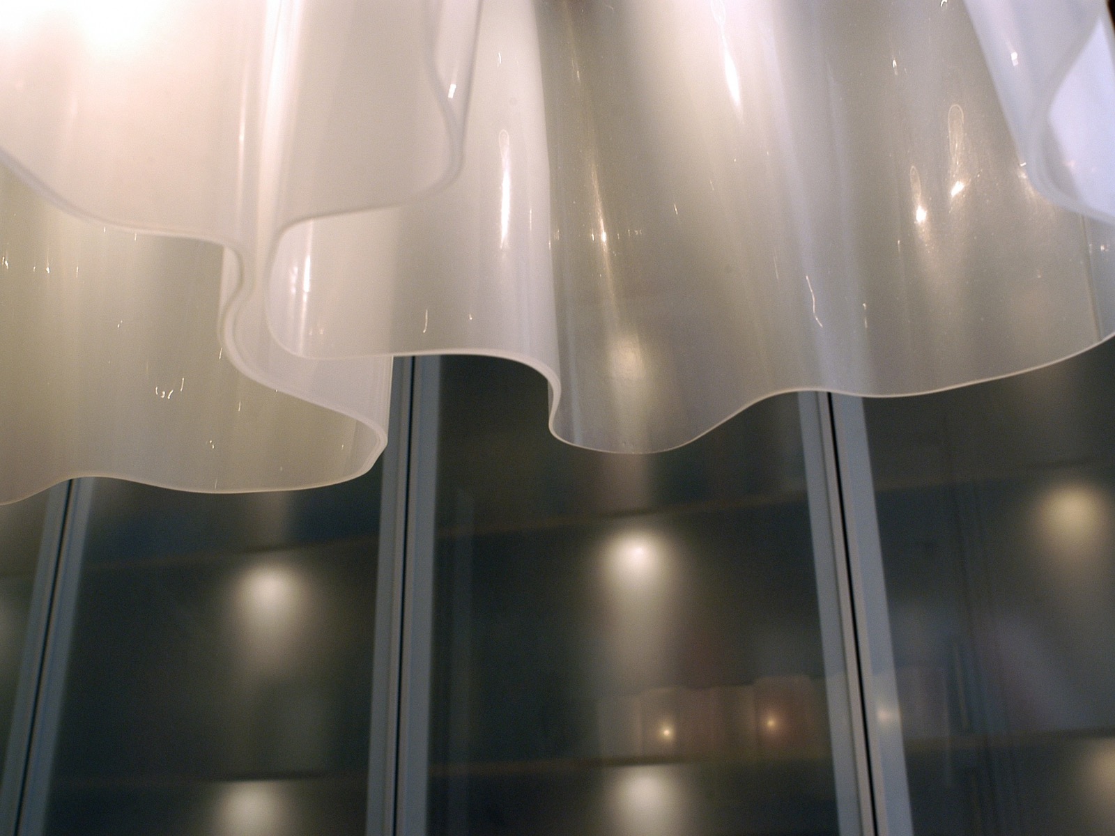 Biała, falista lampa wisząca z tworzywa sztucznego, widziana od dołu, z widocznym fragmentem przeszklonej szafy w tle. Delikatne światło odbija się od powierzchni lampy.