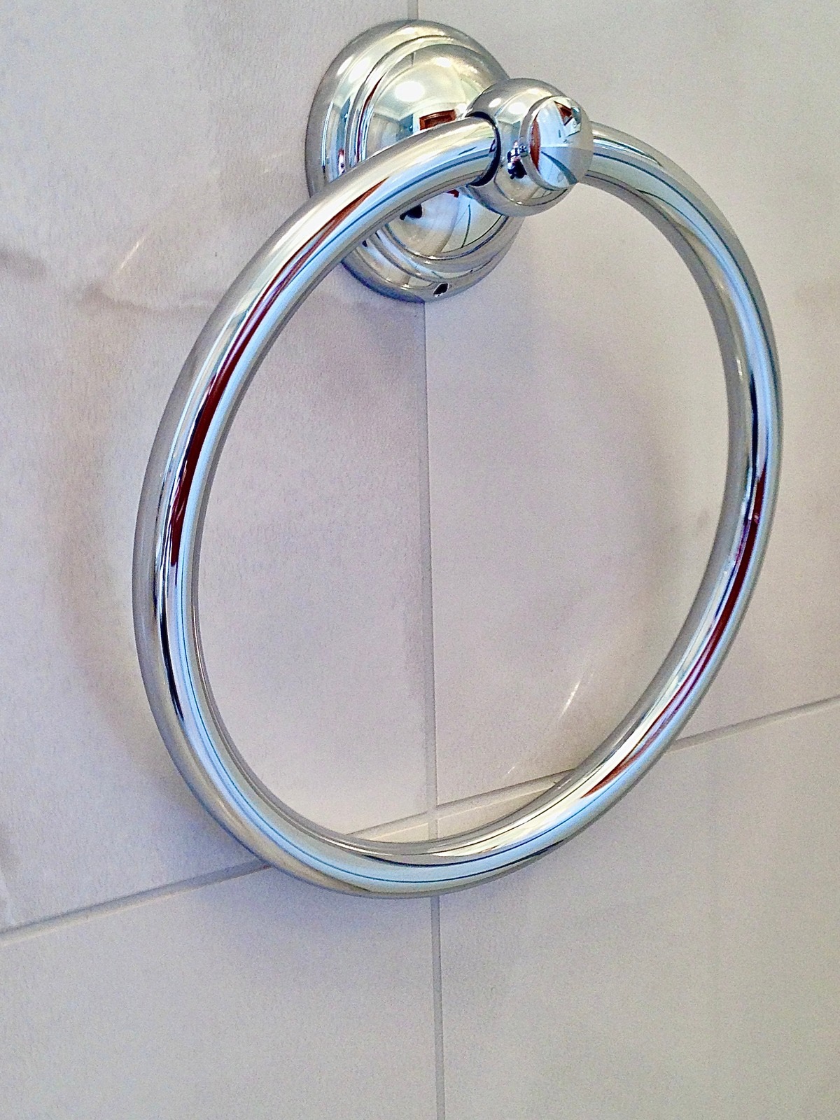 Błyszczący, chromowany wieszak na ręcznik w kształcie koła, zamontowany na białej, kafelkowanej ścianie w łazience. Minimalistyczny design, czyste linie.