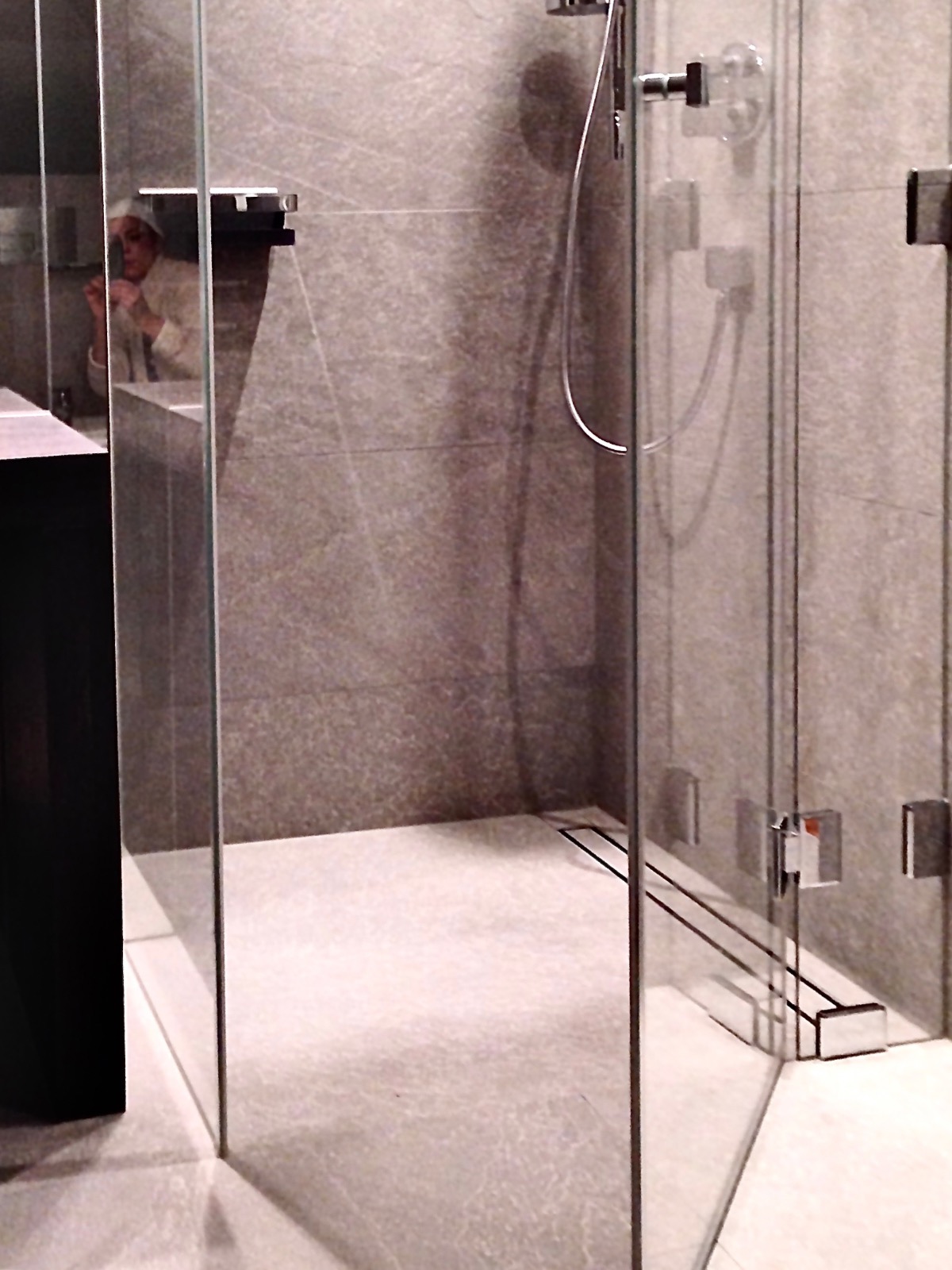 Nowoczesna łazienka z kabiną walk-in, szare płytki i odpływ liniowy. W lustrze odbija się osoba w szlafroku. Minimalistyczny design z akcentem na funkcjonalność.