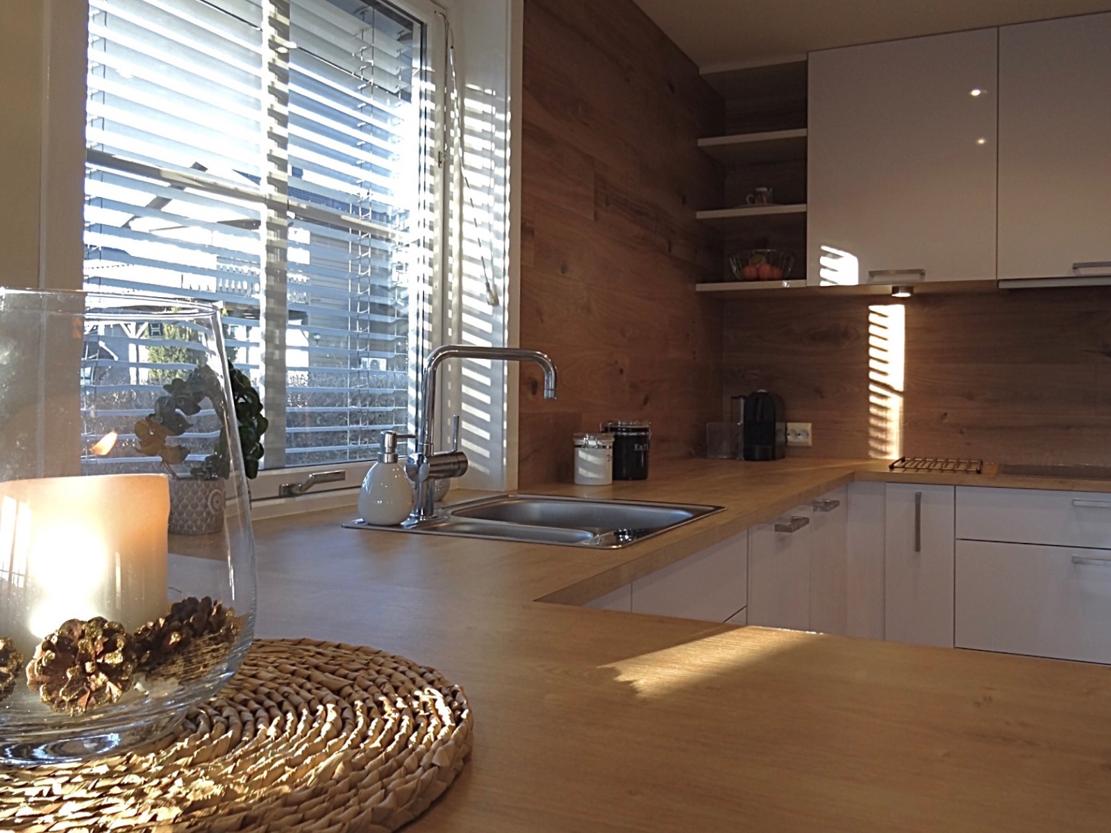 Jasna kuchnia z drewnianymi akcentami: blat, ściana, żaluzje. Ujęcie na zlew, baterię i dekoracje. Białe szafki górne i dolne. Promienie słońca wpadają przez okno.