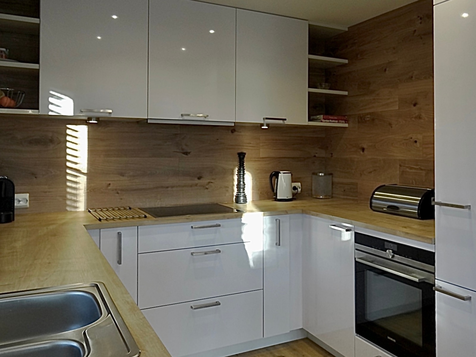 Nowoczesna kuchnia z białymi, lakierowanymi frontami, drewnianym blatem i okładziną ścienną. Widoczny zlew, płyta grzewcza, piekarnik i akcesoria kuchenne.