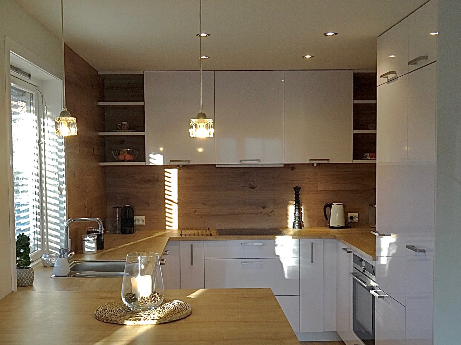 Nowoczesna kuchnia z białymi, błyszczącymi frontami szafek i drewnianym blatem, oświetlona wiszącymi lampami z kryształkami, z dekoracją na blacie w postaci świecy.