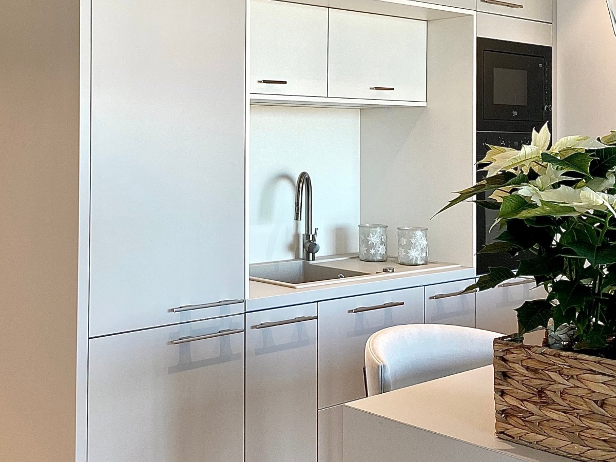 Nowoczesna, biała zabudowa kuchenna z minimalistycznym zlewem i baterią. Na blacie dekoracyjne słoiczki i świąteczna gwiazda betlejemska w plecionym koszu.