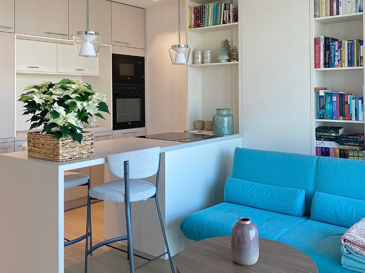 Jasna kuchnia z wyspą i hokerami, połączona z salonem. Na wyspie biała poinsecja w plecionym koszu. W tle zabudowa kuchenna i regał z książkami.